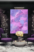 A large vertical abstract wall art print featuring swirling purple, pink, and lilac brushstrokes, displayed in a modern dark living room setting between two purple armchairs and above a round metallic coffee table with a floral arrangement.