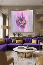 A large square abstract wall art print featuring vibrant purple, pink, and silver brushstrokes, displayed in a modern living room setting above a purple sectional sofa.