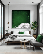 A modern and minimalist bedroom with a large abstract cloud painting on the wall, featuring dark emerald green sky, white clouds and stars. The room has a cozy serene atmosphere, with a low platform bed, dark furniture, and natural elements like a tree branch. A large window overlooking a beach, a gray sofa, and a round gold coffee table Next to it, a sleek gold and chrome minimalist side table holds a coffee cup and a few stylishly arranged books, and there are tall plants in the corners.