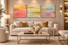 A luxurious living room featuring a three-panel abstract wall art set in pink, yellow, blue purple tones and real gold leaf, hanging over large beige sofa. The room is decorated with gold accents, and floral arrangements. 