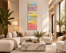 A luxurious living room featuring a three-panel abstract wall art set in pink, yellow, blue purple tones and real gold leaf. The room is decorated with gold accents, and plant arrangements. 
