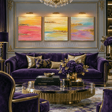 A luxurious living room featuring a purple velvet sofa with gold-trimmed cushions, a round gold marble coffee table, and a three-panel abstract wall art set in pink, yellow, and blue tones. The room is decorated with gold accents, crystal chandeliers, and purple floral arrangements.