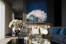 A large square canvas wall art print depicting a dramatic blue sky with a glowing white cloud, displayed in a modern living room setting above a fireplace.