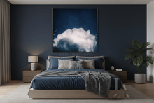 A modern bedroom featuring a large framed square wall art print by Julia Apostolova depicting a dramatic white cloud against a deep navy blue night sky. The artwork is mounted on a dark navy blue accent wall above a bed with blue and gray bedding.