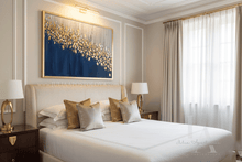 Neatly made bed in a bedroom with a large abstract painting on the wall with gold leaf, navy blue and silver/ gray colors, by artist Julia Apostolova, decorative gold silver pillowa, lamps, and curtains.