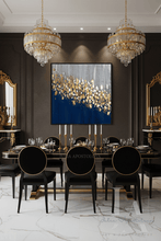 A luxury dining room featuring a large square original abstract wall art with a deep blue background and shimmering gold leaf like-flowers pattern, hanging above a black dining table and chairs with gold trim.