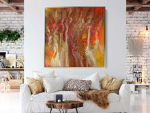 Abstract artwork featuring swirling patterns in shades of copper, orange, and gold, displayed on a white wall above a cozy white sofa adorned with decorative pillows and a brown throw. A wooden coffee table with a small black object sits in front of the sofa. The background includes a light-colored brick wall .