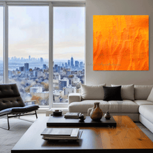 A large abstract orange painting on the wall , displayed in a modern living room with a large window overlooking a cityscape .  The room has a neutral color scheme with a large light gray sofa, gray and black pillows, a low wooden coffee table, and a gray rug on the floor.