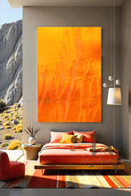 A modern and minimalist bedroom with a large orange abstract painting on the wall , a bed with orange sheets and orange and yellow pillows . The room has a cozy serene atmosphere, with a low platform bed, orange poof and wooden furniture, and natural elements like a vases , lamps , and a large window overlooking a natural rocky landscape .