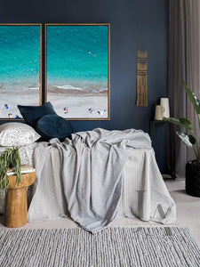 A large coastal-inspired minimalist beach wall art set on the wall, over bed, in boho bedroom decor setting. The room features dark gray walls, a light gray woolen carpet, bed surrounded with pillows, plans, a low wooden coffee table and a decorative boho accents. The coastal Aerial wall art canvas set futures, people on the beach, ocean waves, and turquoise waters, making an relaxing, cosy and calming atmosphere.

