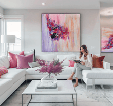 A woman sits on a white sectional sofa in a bright modern living room, reading a book. The room features a large abstract painting in pink, purple, lilac, and peach tones hanging on the wall above the sofa.