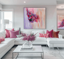 A large square abstract canvas painting with vibrant pink, purple, lilac, and peach brushstrokes hangs on a light gray wall above a modern white sectional sofa decorated with pink and magenta pillows.