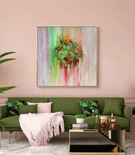 A square framed abstract painting with a green floral wreath motif hangs on a dusty pink wall above a green sofa in a styled living room.