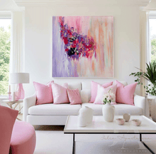 A large abstract canvas painting with vibrant pink, purple, and peach brushstrokes hangs on a white wall above a modern white sofa styled with pink pillows in a bright, contemporary living room.