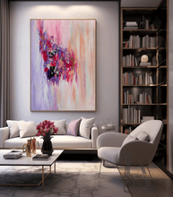 A large abstract canvas painting with vibrant pink, purple, red, and peach brushstrokes hangs on a light gray wall in a modern living room setting with a white sofa, a gray armchair, and a tall bookshelf.
