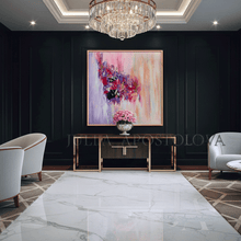 A large abstract canvas painting with pink, purple, lilac, and peach brushstrokes hangs on a dark paneled wall in a luxurious modern living room. The room features a white marble floor, a gold-framed console table with a pink flower arrangement, two white armchairs, and a large crystal chandelier overhead.