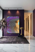 A large framed abstract wall art print hangs on a dark purple brick wall in a styled interior. The artwork features swirling purple, lavender, peach, cream, and gold tones with a textured cracked pattern.