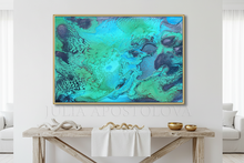 An abstract framed wall art print featuring swirling teal, turquoise, green, and dark blue marbled patterns with a thin gold frame, displayed above a rustic wooden console table in a bright, modern living room.
