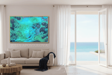 A large framed abstract painting with swirling turquoise, teal, and dark blue-green patterns hangs on a beige wall above a light beige sofa in a bright modern living room. Large white curtains frame an open glass door overlooking a blue ocean and white sand beach.