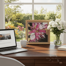Desk with laptop, framed artwork, mug, and flowers in a vase near a window with a garden view.