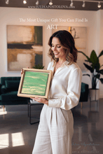 A woman in a white blouse and white pants stands in a modern art gallery or studio, holding a small framed painting with green and gold brushstrokes. The background features large abstract paintings, a dark green velvet sofa, and potted plants.