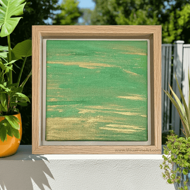 Framed abstract artwork with green and gold hues on a white surface, surrounded by plants.