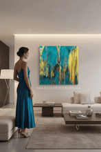 Woman in a teal dress standing in a modern living room with a colorful teal gold abstract painting on the wall. The painting is created by artist Julia Apostolova and futures gold leaf accents.