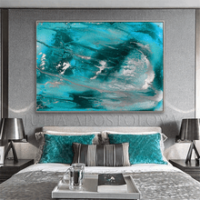 A large square abstract wall art print with swirling turquoise, teal, aqua, and silver brushstrokes hanging above a neatly made bed in a modern gray bedroom. The artwork is the product being shown, but because it is displayed in a real interior environment with bed, lamps, and nightstands visible, this fits Lifestyle rather than Product/Packaging.