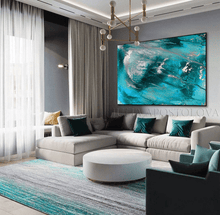 A large square abstract canvas print featuring vibrant turquoise, teal, and silver waves, displayed on a gray wall above a modern light gray sectional sofa in a contemporary living room.