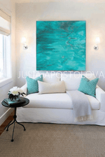 A large square abstract painting in vibrant turquoise and teal hangs on a white wall above a white sofa with decorative pillows and a throw blanket in a bright living room.