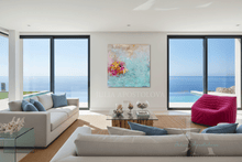 An abstract coastal painting with turquoise, pink, and gold tones hangs on a white wall in a modern, sunlit living room with ocean views, a white sofa, and a bright magenta chair.