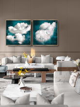 A modern living room featuring a pair of square framed wall art prints depicting fluffy white clouds against a deep Transformative teal sky, hung on a brown paneled wall above a light gray sofa.