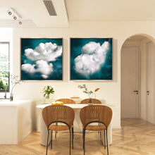 Two framed square wall art prints showing large white clouds against a deep teal-blue sky, displayed above a dining table in a bright modern interior with chairs, flowers, and an arched doorway.