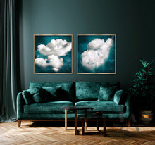 A modern living room scene featuring two framed square wall art prints with white clouds against a deep teal sky, hung above a dark teal velvet sofa. The room has a dark teal wall, wood herringbone flooring, a dark curtain on the left, and a potted plant on the right.