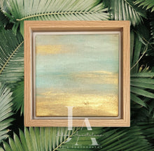 A framed original painting depicting a serene landscape with a sunset sky. The painting shows a tranquil scene with a warm, golden horizon and soft, pastel colors in the sky. The frame is made of light-colored wood, adding to the natural, calming feel of the artwork, surrounded by lush green palm leaves.