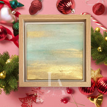 A wooden frame with a painting of a sunset landscape with metallic gold and pale turquoise color schemes, surrounded by Christmas ornaments and greenery on a pink background.