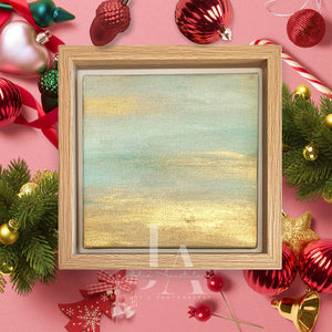 A wooden frame with a painting of a sunset landscape with metallic gold and pale turquoise color schemes, surrounded by Christmas ornaments and greenery on a pink background.