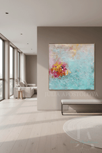 An abstract canvas painting with a turquoise blue background and a colorful floral-like burst in pink, yellow, and orange, displayed on a beige wall in a modern, sunlit living room. The text 'JULIA APOSTOLOVA' is overlaid near the bottom.
