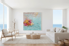 A large abstract canvas painting with turquoise, pink, yellow, and gold brushstrokes hangs on a white wall in a bright, modern living room with a light gray sofa, wooden coffee table, and ocean views through the windows.