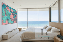 A modern bedroom with a large abstract wall art piece featuring teal, pink, and green marbled colors, positioned next to a bed with beige linens and a woven headboard, overlooking a beach and ocean view.