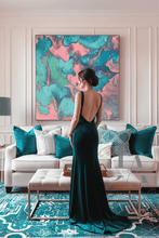 A woman stands in a living room wearing a floor-length dark teal gown with a low open back. The room features a white sofa with teal and white pillows, a large abstract painting on the wall in turquoise, teal, and pink tones, and a patterned teal rug.