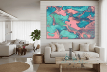 A large square abstract canvas print with swirling teal, turquoise, and pink paint patterns, displayed on a white shiplap wall above a modern beige sofa in a bright, contemporary living room.