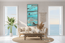 A vertical abstract canvas print by artist Julia Apostolova , with turquoise, teal, gold, and gray marbled patterns hangs on a light gray wall in a bright, modern living room. The room features a beige sofa, a wooden coffee table, a woven jute rug, a potted plant, and a macrame wall hanging, with large windows looking out to the ocean.
