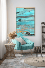 A framed abstract canvas print by artist Julia Apostolova with swirling turquoise, teal, gray, and black paint patterns hangs on a light gray wall in a styled living room setting with a teal velvet armchair, a wicker side table, and a round rug.