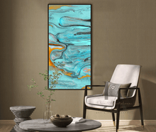 A framed abstract canvas print by artist Julia Apostolova, with swirling turquoise, blue, gray, and gold paint patterns, displayed on a textured beige wall in a modern living room setting next to a white armchair.