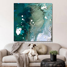 A large abstract canvas wall art print with teal, green, white, and blue marbled swirls hanging above a light beige sofa in a living room. A white and black dog is lying on the sofa, with a round beige pillow and a gray knit throw blanket nearby.