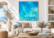 A large square abstract canvas painting with vibrant turquoise, blue, and white watercolor-like textures hangs on a white wall above a beige sofa in a bright, modern coastal-style living room.