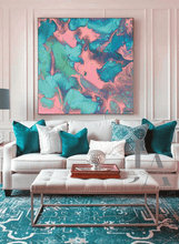 A large square abstract wall art print with swirling teal, turquoise, pink, and cream paint patterns, displayed above a white sofa in a styled living room.