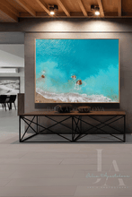 A large framed square wall art print depicting turquoise ocean water with white surf and small shells, displayed above a wooden console table in a modern interior with wood ceiling beams and track lighting.