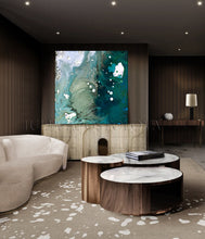 A large abstract wall art piece with swirling teal, blue, green, and white paint is displayed above a console table in a modern living room with dark wood slat walls, a curved cream sofa, and round marble-top coffee tables.